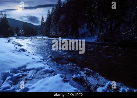 Tereblya Fluss im Winter bei Vollmondlicht. Schneebedeckte Küste mit Nadelwald am Ufer. Berge unter einem bewölkten Himmel in frostiger Nacht Stockfoto