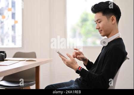 Hübscher junger asiatischer Büroangestellter oder Geschäftsmann im lässigen Outfit sitzt an seinem Schreibtisch, nutzt eine Anwendung auf seinem Smartphone. Rückansicht Stockfoto