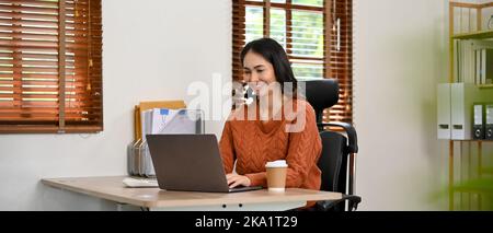 Charmante, tausendjährige asiatische Geschäftsfrau oder Sekretärin, die an ihrem Schreibtisch mit einem Laptop arbeitet. Stockfoto