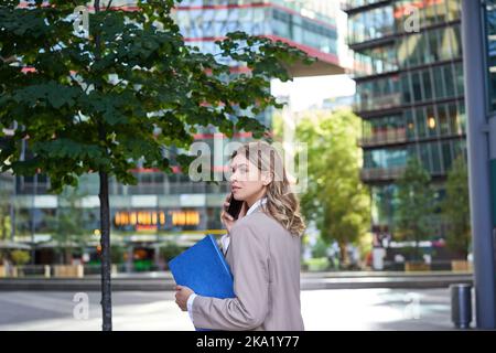 Porträt einer Frau, die beim Gehen zum Bürogebäude einen Anruf entgegennimmt. Geschäftsfrau, die jemanden anruft, Dokumente hält Stockfoto