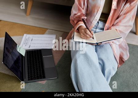 Nahaufnahme einer jungen Schülerin in Casualwear, die auf dem Boden sitzt und sich während des Online-Unterrichts mit der Tutorin Notizen in einem Notizbuch macht Stockfoto