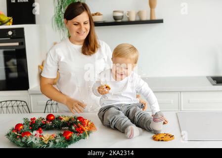 Vorbereitung auf Weihnachten. Mein Sohn sitzt in der Küche auf dem Tisch und meine Mutter hält Weihnachtsdekorationen. Das Kind lacht Stockfoto