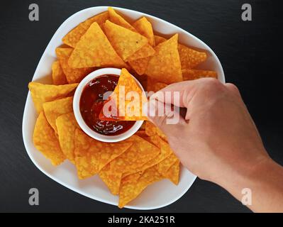Männliche Hand taucht einen mexikanischen Maischip oder Tortilla Chip in einen würzigen Salsa Dip, Draufsicht auf eine Schüssel mit Tortillas mit Salsa auf schwarzem Schiefer Hintergrund Stockfoto
