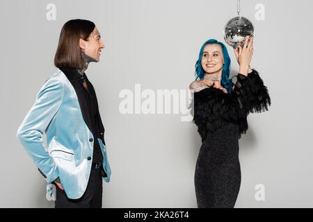 Lächelnde queere Person in blauer Perücke berühren Disco-Ball in der Nähe von eleganten nicht binären Freund auf grauem Hintergrund, Stockbild Stockfoto