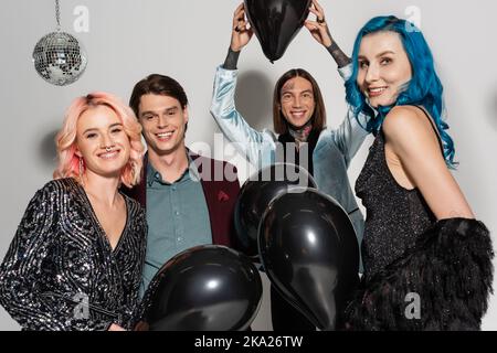 Fröhliche queere Person hält schwarzen Ballon über Kopf in der Nähe von nicht binären Freunden lächeln an der Kamera auf grauem Hintergrund, Stock Bild Stockfoto