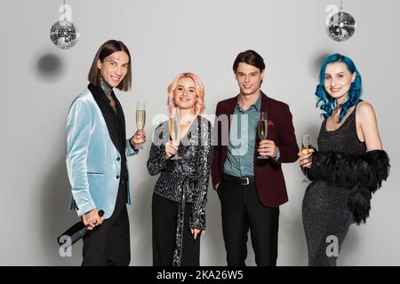 Lächelnde nichtbinäre Freunde in eleganten festlichen Kleidern halten Champagner während Neujahr Party auf grauem Hintergrund, Stockbild Stockfoto