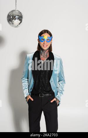 Fröhliche tätowierte nichtbinäre Person in Party-Maske stehend mit Händen in Taschen auf grauem Hintergrund, Stockbild Stockfoto