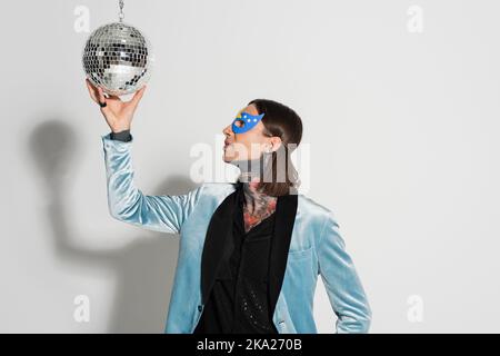 Queer Person in eleganten Seide Blazer und Party-Maske berühren glänzenden Disco Ball auf grauem Hintergrund, Stock Bild Stockfoto