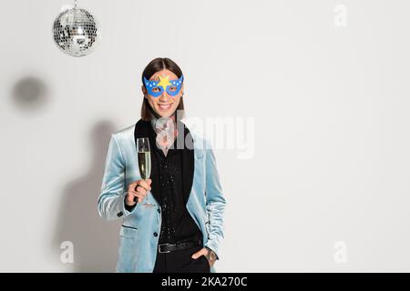 Stilvolle queere Person in Party-Maske stehen mit Hand in der Tasche und Champagner-Glas in der Nähe Disco Ball auf grauem Hintergrund, Stock Bild Stockfoto