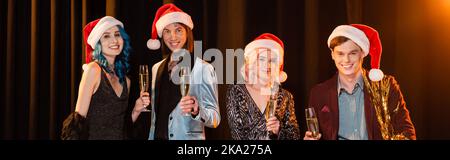 Elegante nichtbinäre Freunde in weihnachtsmützen lächeln der Kamera zu, während sie Champagner auf dunklem Hintergrund mit Beleuchtung, Banner, Stockbild halten Stockfoto