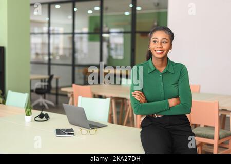 Erfolgreiche junge afroamerikanische Unternehmerin, Kleinunternehmerin, Büroangestellte, schwarze Geschäftsfrau mit grünem Casual-Hemd steht in selbstbewusster Pose mit gekreuzten Armen Stockfoto