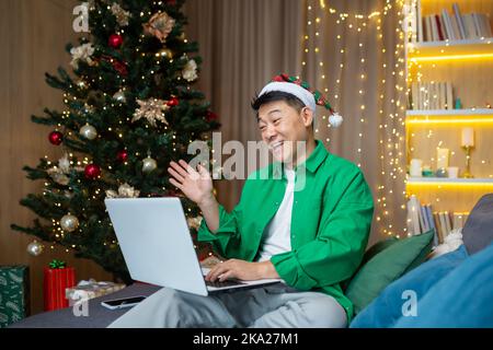 Ein glücklicher junger Mann in einem grünen Hemd und einem roten Weihnachtsmannhut sitzt auf einem Sofa in der Nähe eines Weihnachtsbaums und hält einen Laptop. Kommuniziert vor der Kamera und wünscht Familie und Freunden frohe Feiertage. Stockfoto