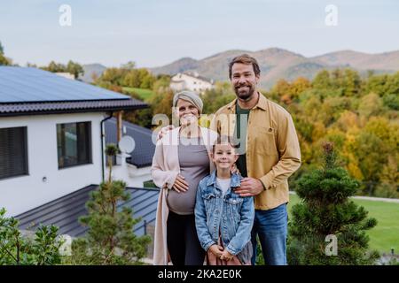 Glückliche Familie in der Nähe ihres Hauses mit Sonnenkollektoren. Alternative Energie, Ressourcenschonung und nachhaltiges Lifestyle-Konzept. Stockfoto