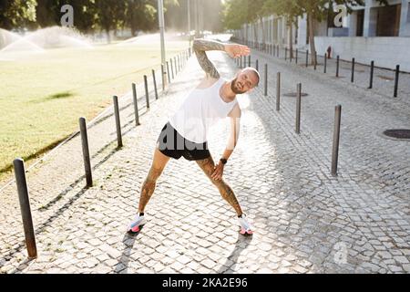 Athletischer junger tätowierte Mann, der sich vor den Übungen im Freien durch Dehnen aufwärmt, bärtiger, falter Mann in Sportswear, der Trainingsübungen macht Stockfoto