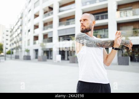 Porträt eines männlichen Athleten, der bei Sonnenuntergang die Arme im Freien streckt, bärtig tätowierte Glatze, die sich vor dem Training unter freiem Himmel aufwärmt Stockfoto