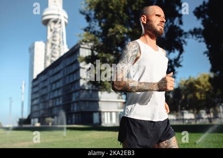 Seitenansicht des athletischen bärtigen tätowierten kaukasischen Mannes, der morgens draußen in der Stadt joggt, gesunder, athletischer männlicher Sportler, der läuft Stockfoto