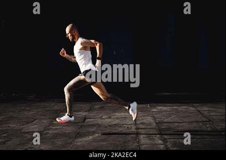 Kahlköpfiger junger athletischer tätowierte Mann läuft schnell im Freien, Sportler bereitet sich auf den Marathon vor, macht Cardio-Übungen im Freien, Seitenansicht Stockfoto