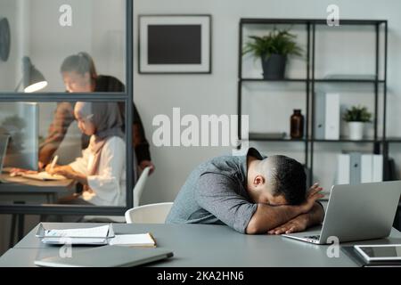 Ein junger erschöpfter Mann, der den Kopf auf dem Schreibtisch hält, während er vor dem Laptop sitzt und zwei weibliche Kollegen im Büro sieht Stockfoto