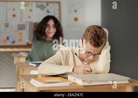 Selektiver Fokus auf clevere und fleißige Schüler Notizen im Copybook in der Lektion gegen grausame Klassenkamerad lachend ihn Stockfoto