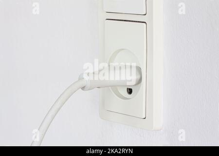 Nahaufnahme des Steckers eines elektrischen Geräts, das an eine Steckdose auf einem weißen Wandhintergrund angeschlossen ist. Steckdose weiß mit Kabel. Strom Stockfoto