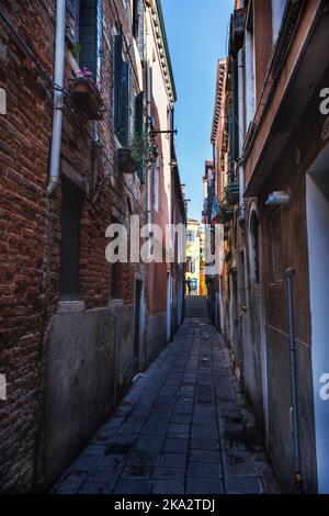 Dunkle und schmale Gasse in Venedig, die zu sonnigen Häusern führt Stockfoto