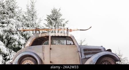 Verwitterter Oldtimer mit Skiern, die an einem Dachträger vor schneebedeckten Tannen befestigt sind Stockfoto