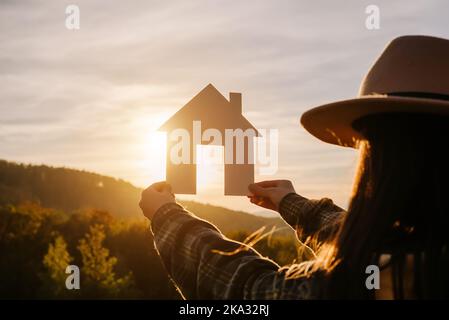 Seitenansicht der weiblichen Hände, die ein kleines Papierhaus vor Sonnenuntergang- oder Sonnenaufgangslicht auf dem Hintergrund der Berge halten. Zuhause in Sonnenstrahlen in der herbstlichen Natur. Fam Stockfoto