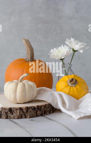 3 Kürbisse auf einem Holzblock, Milchflasche mit 2 weißen Chrysanthemen und cremefarbene Leinenserviette vor hellem Hintergrund. Mit Platz für Kopien Stockfoto