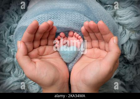 Die Handflächen des Vaters, die Mutter halten die Beine des Neugeborenen. Der Fuß des Kindes auf den Handflächen der Eltern mit einem blau gestrickten Herz auf den Fersen. Stockfoto