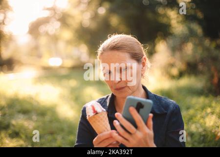 Reife Frau mit Smartphone und Eis im Park Stockfoto
