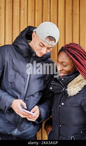 Lächelndes multiethnisches Paar, das auf der Straße auf das Handy schaut. Stockfoto
