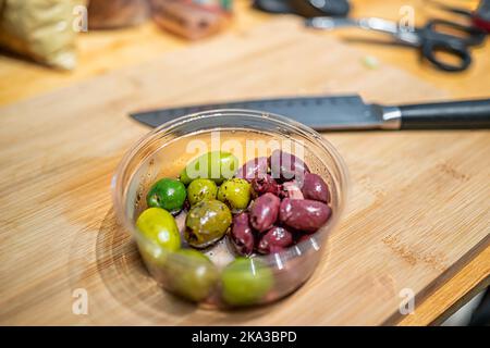 Bambusschneidebrett mit Messer und Nahaufnahme von sortierten grünen und kalamata-Oliven in Plastikbehältern von Lebensmittelgeschäft, Barbuffet und Marinebrot Stockfoto