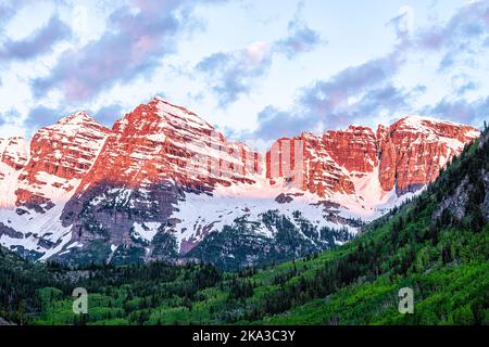 Kastanienbraune Glocken Gipfeln aus der Nähe mit rotem Sonnenaufgangslicht auf schneebedeckten felsigen Bergen in Aspen, Colorado, mit blauem, bewölktem Himmel im Juni, Frühsommer dra Stockfoto