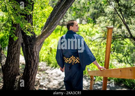 Junger, fröhlich lächelnder Mann im Kimono-Kostüm, der sich im japanischen Garten mit Blick auf die Natur auf einen Geländerzaun stützt und den Kopf dreht Stockfoto
