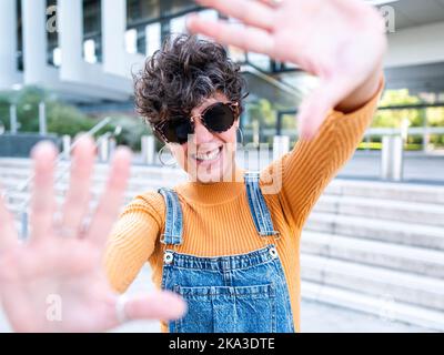 Fröhliche ethnische Frau in Denim-Overall und Sonnenbrille mit Fotogesten, während sie auf der Straße in Sonnenschein gegen ein modernes Gebäude steht Stockfoto