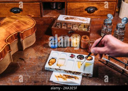 Hoher Winkel des gesichtslosen Geigenbauers, der am Tisch in eigener Geigenbauerei arbeitet und perfekte Holzfarbe für Violininstrument schafft Stockfoto