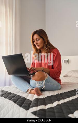 Barfuß hispanische Frau in legerer Kleidung sitzend mit gekreuzten Beinen auf dem Bett und im hellen Schlafzimmer zu Hause im Netbook surfen Stockfoto