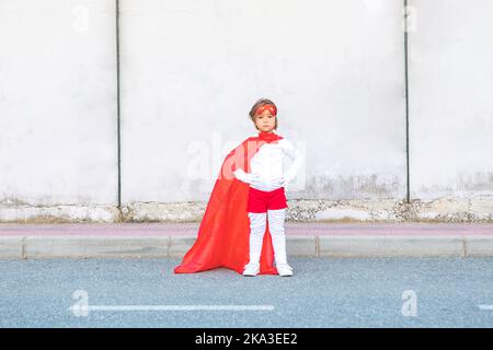 Niedliches kleines Mädchen im Superhelden-Kostüm und rotem Umhang, das mit den Händen an der Taille gegen die Betonwand steht Stockfoto