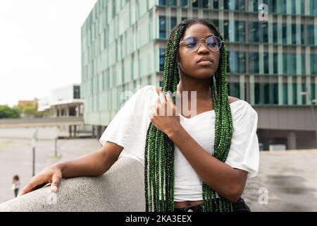 Junge, coole afroamerikanische Frau in weißem Top mit langen geflochtenen Haaren, die eine Brille trägt und selbstbewusst in die Stadt schaut Stockfoto