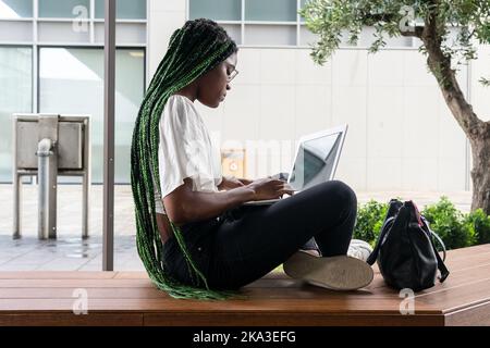 Seitenansicht einer jungen afroamerikanischen Freiberuflerin mit langen geflochtenen Haaren und in einer Brille, die auf einer Holzbank sitzt und in der Stadt auf einem Laptop herumstöbert Stockfoto