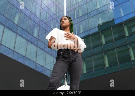 Von unten zeigt eine coole junge afroamerikanische Frau in weißem Top und Denim mit langen Zöpfen, die in der Nähe eines modernen Hochhauses stehen und wegschauen Stockfoto