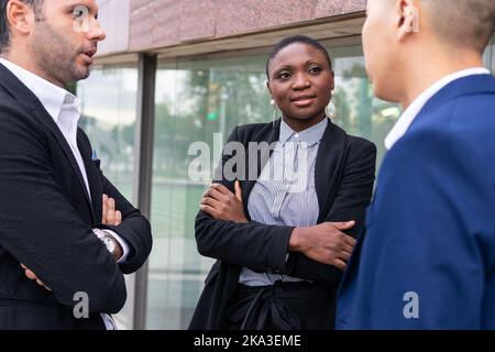Positive afroamerikanische Frau in formellem Anzug, die mit gekreuzten Armen an einer Glaswand steht und mit männlichen Kollegen über die Geschäftsstrategie diskutiert Stockfoto