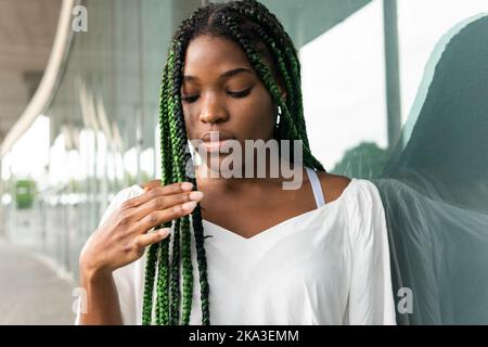 Coole junge afroamerikanische Frau mit langen Afrogeflechten, die an einer reflektierenden Straßenwand stehen und nach unten schauen Stockfoto
