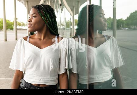Coole junge afroamerikanische Frau mit langen Afrogeflechten, die an einer reflektierenden Straßenwand stehen und wegschauen Stockfoto