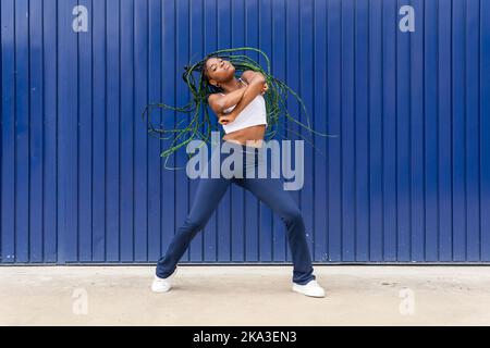 Coole afroamerikanische Frauen mit vollem Körper in Denim und weißem Oberteil mit langen zitternden Zöpfen, die auf dem Bürgersteig gegen die blaue Metallwand Freestyle tanzen Stockfoto