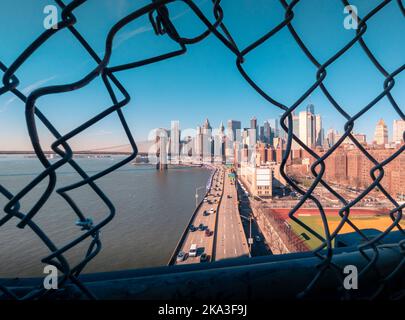 FDR Drive mit Autos in der Nähe der Brooklyn Bridge am Ufer des East River gegen wolkenlosen Sonnenuntergang Himmel hinter Loch im Netzzaun in Manhattan, New York, Stockfoto