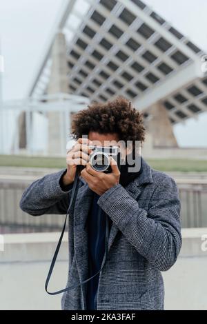 Afroamerikanischer Mann mit Afro-Frisur, der auf der Straße mit einer Vintage-Fotokamera fotografiert Stockfoto