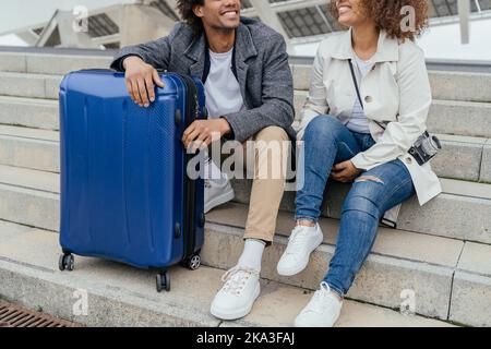 Ein heiteres afroamerikanisches Paar in legerer Kleidung sitzt auf einer Treppe und schaut während des Urlaubs in die Stadt Stockfoto