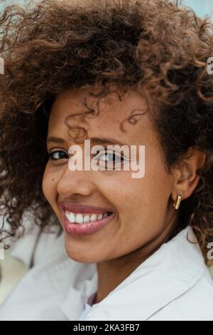 Glückliche junge afroamerikanische Frau mit lockigen Haaren lächelt und schaut tagsüber auf die Kamera Stockfoto