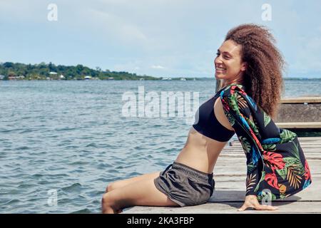Seitenansicht einer fröhlichen ethnischen Frau mit lockigen Haaren, die Shorts mit Oberteil und Hemd auf einem schäbigen Holzsteg mit Beinen in klarem blauem Wasser trägt Stockfoto
